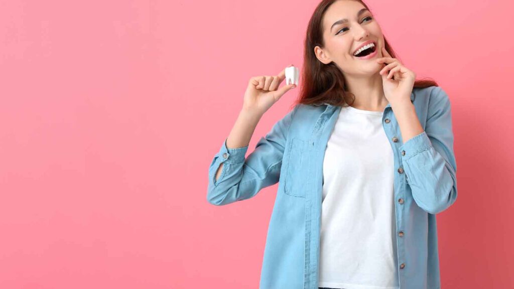 A smiling woman in a blue shirt holding a tooth model against a pink background